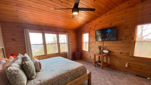 Ein Schlafzimmer einer Holzhütte mit Bett, Deckenventilator, Fernseher und großen Fenstern.