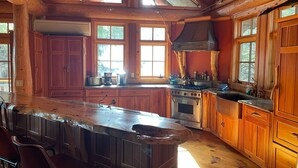 A rustic kitchen with wooden cabinets, a large island with bar stools, a stove, and a window overlooking greenery. - Cayuga Lodge  (Cooperstown)
