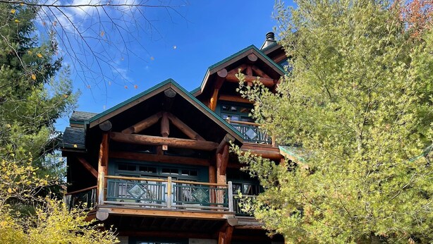 A multi-story log cabin with a balcony, stone steps, and a landscaped garden with rocks and shrubs. - Cayuga Lodge  (Cooperstown)