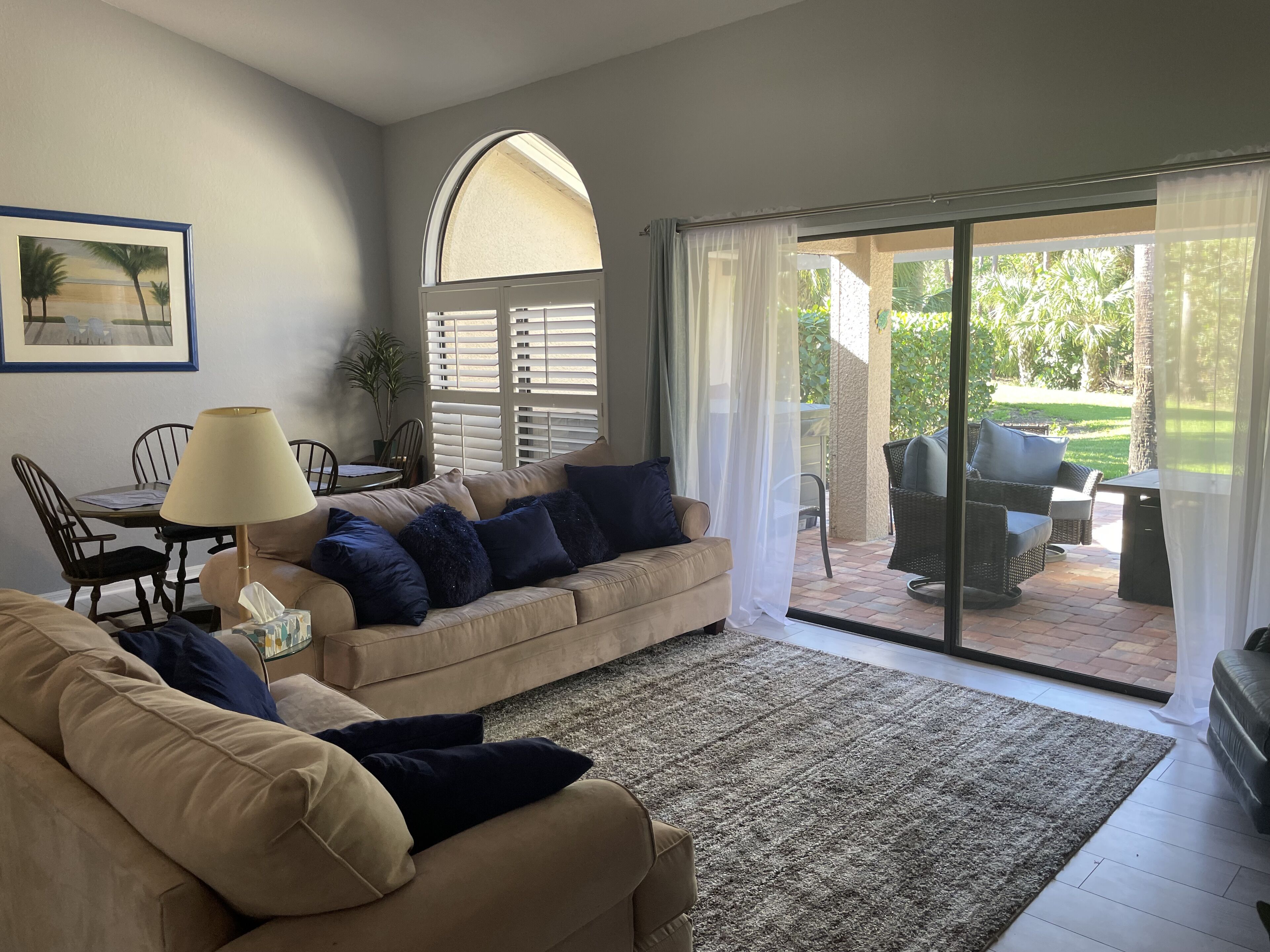 A living room with a beige sofa, a table with chairs, a lamp, and a view of an outdoor patio area with wicker furniture and a glass door.