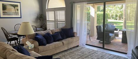 A living room with a beige sofa, a table with chairs, a lamp, and a view of an outdoor patio area with wicker furniture and a glass door.