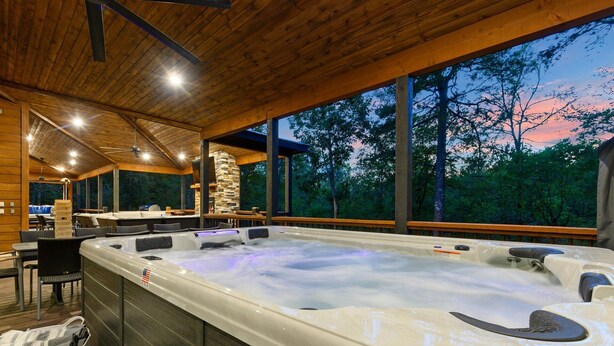 Un jacuzzi en un área exterior techada con vigas de madera, paredes de piedra y vista a los árboles al anochecer.
