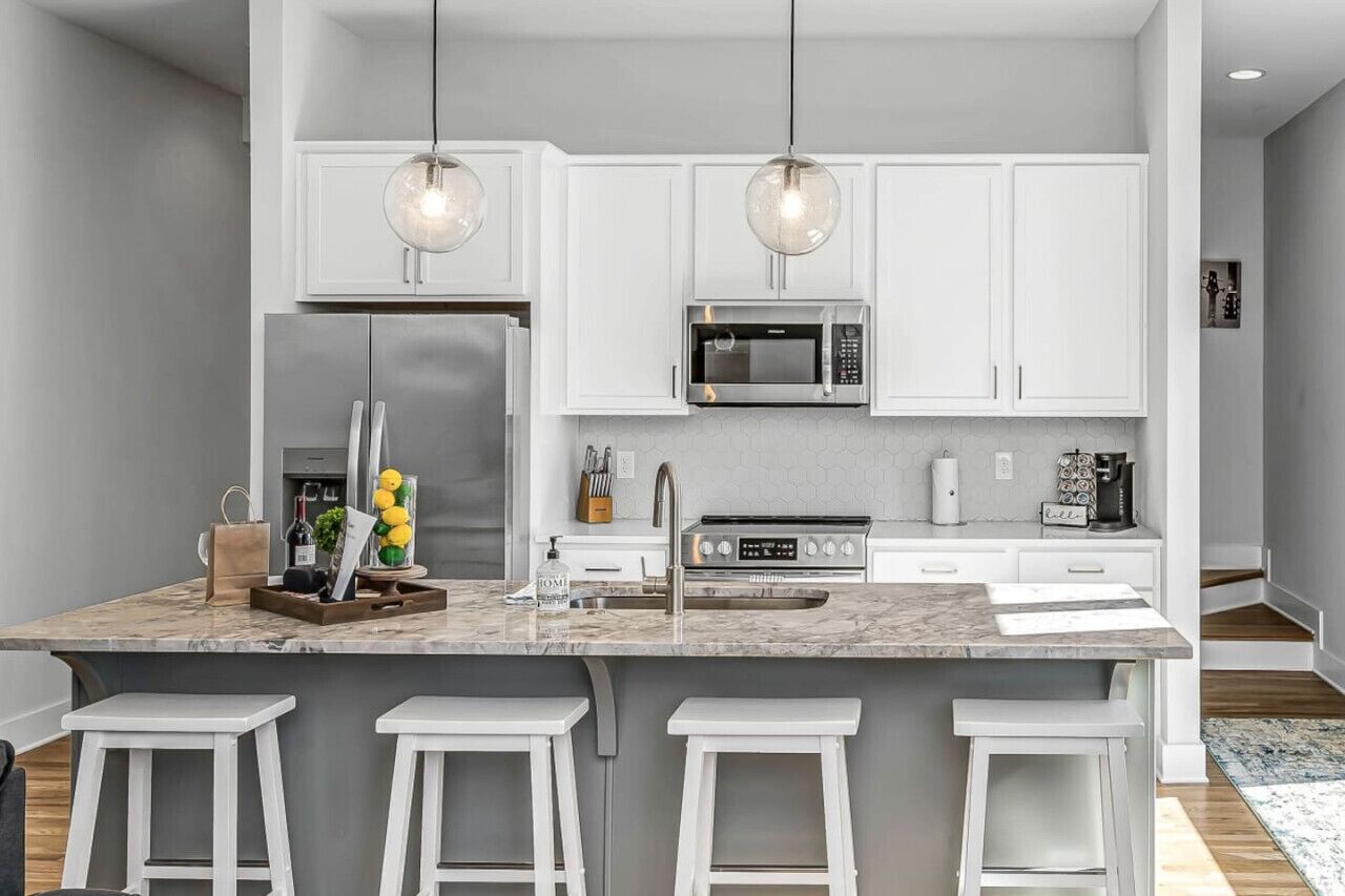 A modern kitchen with white cabinets, a stainless steel refrigerator, a microwave, and a gas stove. There are three white bar stools and a marble countertop.