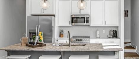 A modern kitchen with white cabinets, a stainless steel refrigerator, a microwave, and a gas stove. There are three white bar stools and a marble countertop.