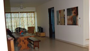 Ein geräumiges Wohnzimmer mit Esstisch aus Holz, einer Couch und einem Fernseher. An der Wand hängen drei gerahmte Bilder und es gibt einen Deckenventilator.