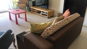Ein Wohnzimmer mit braunem Sofa, einem roten Tisch, einem Fernseher auf einem Holzgestell und einem gemusterten Kissen.