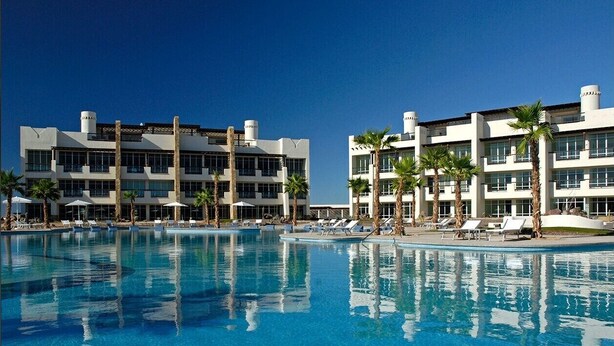 Ein großer Pool mit Liegestühlen und Sonnenschirmen, umgeben von modernen Gebäuden mit mehreren Etagen und großen Fenstern.