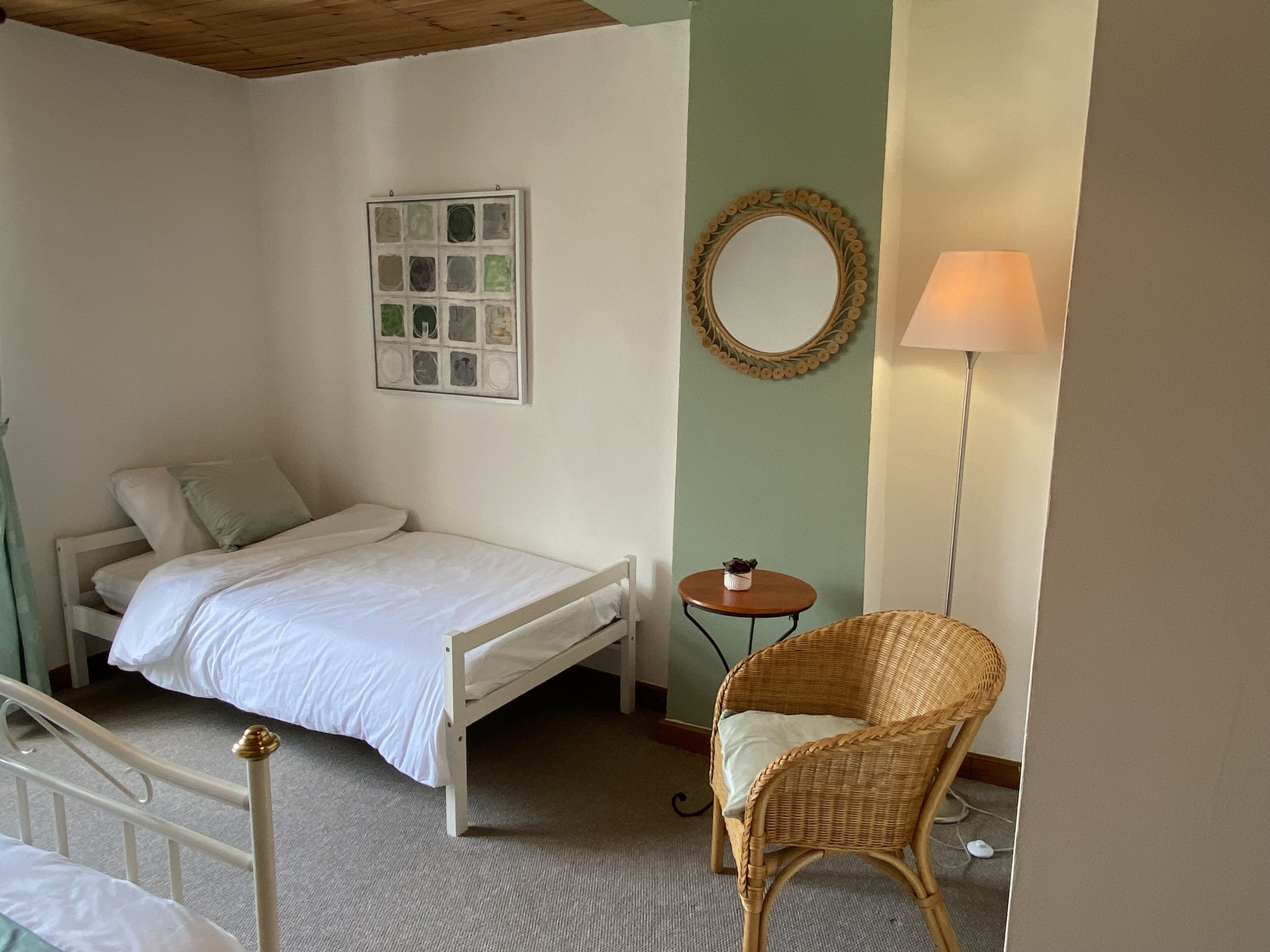 A bedroom with a single bed, a small round table, a wicker chair, a wall-mounted mirror, and a lamp.