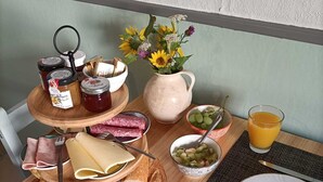 Een ontbijtsetting met croissants, kaas, ham, broodjes, een fruitschaal, een vaas met bloemen, een glas sinaasappelsap en toevoegingen op een houten tafel.