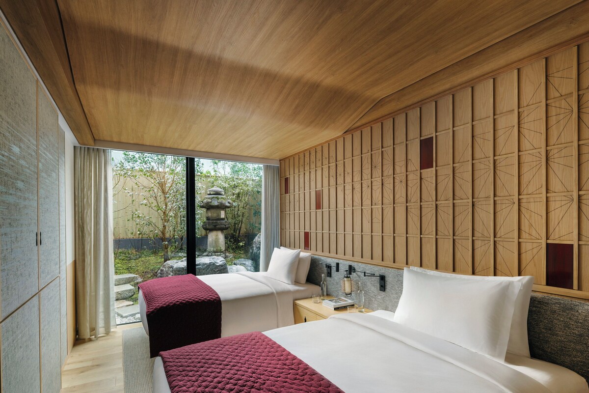 A modern hotel room with two beds, a wooden ceiling, a view of a garden, and a traditional Japanese-style wall panel.