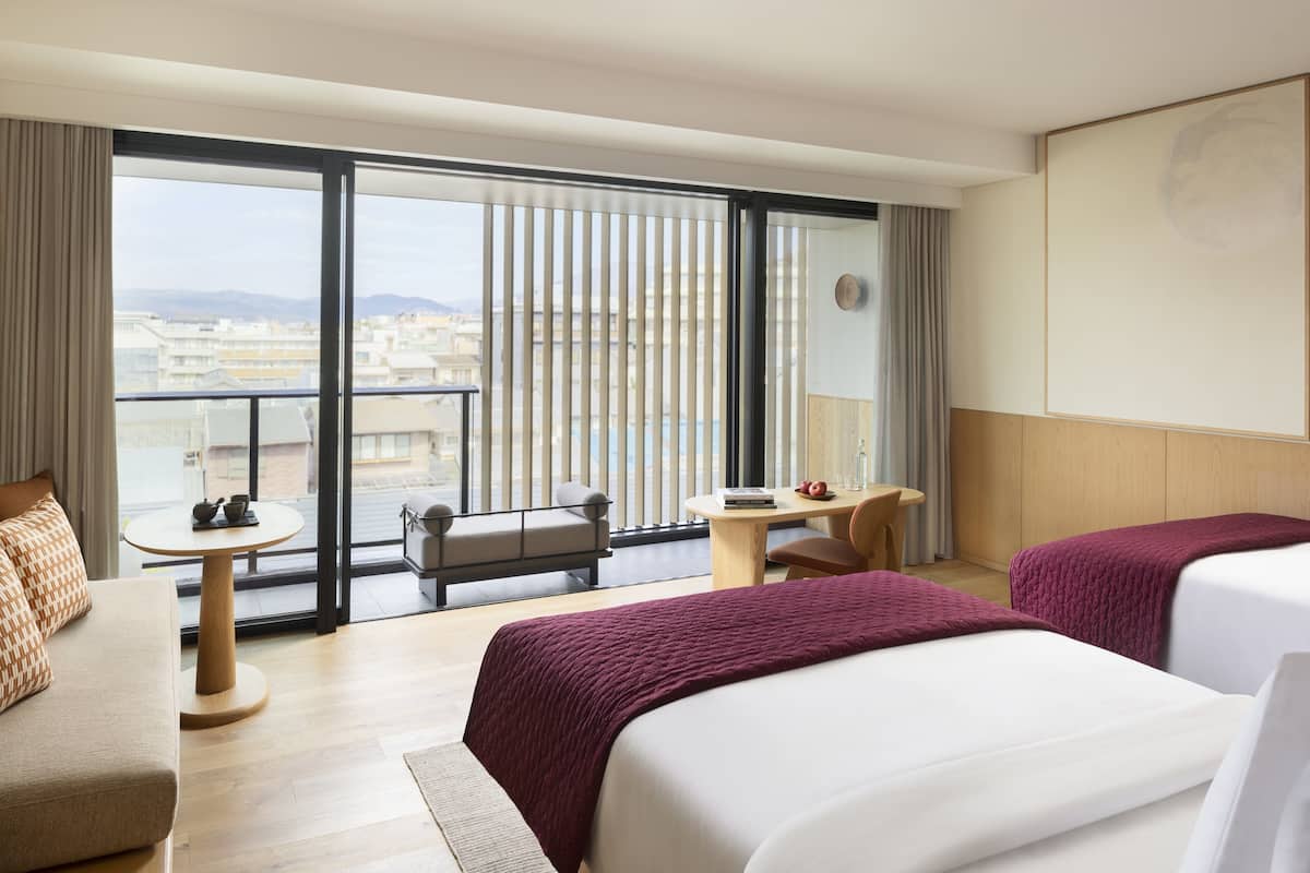A hotel room with two beds, a sofa, a small table, and a balcony with a view of buildings and mountains.