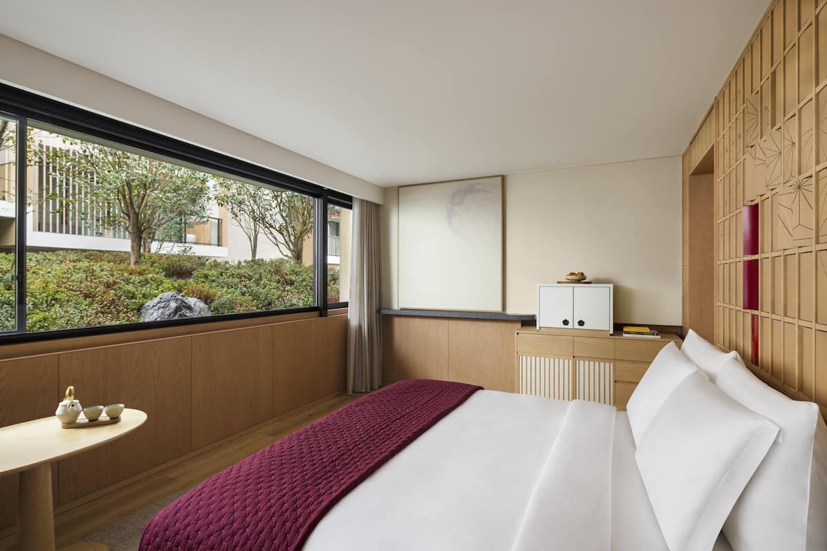 A modern hotel room with a large window, a bed, a small table with tea set, and a cabinet.