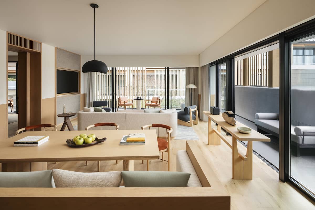 A modern living room with a sofa, dining table, and chairs. There is a large window with blinds, a hanging light fixture, and a view of the outside.