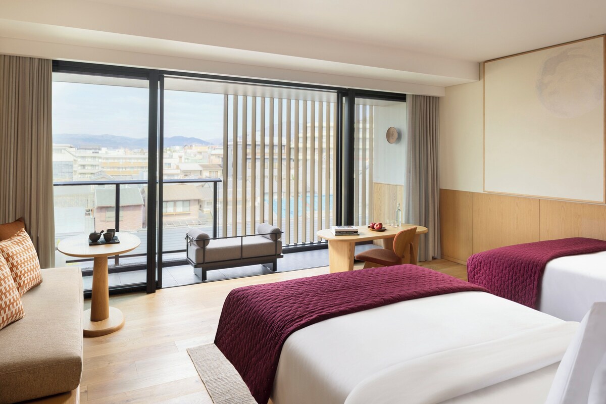 A hotel room with two beds, a sofa, a small table, and a balcony with a view of buildings and mountains.