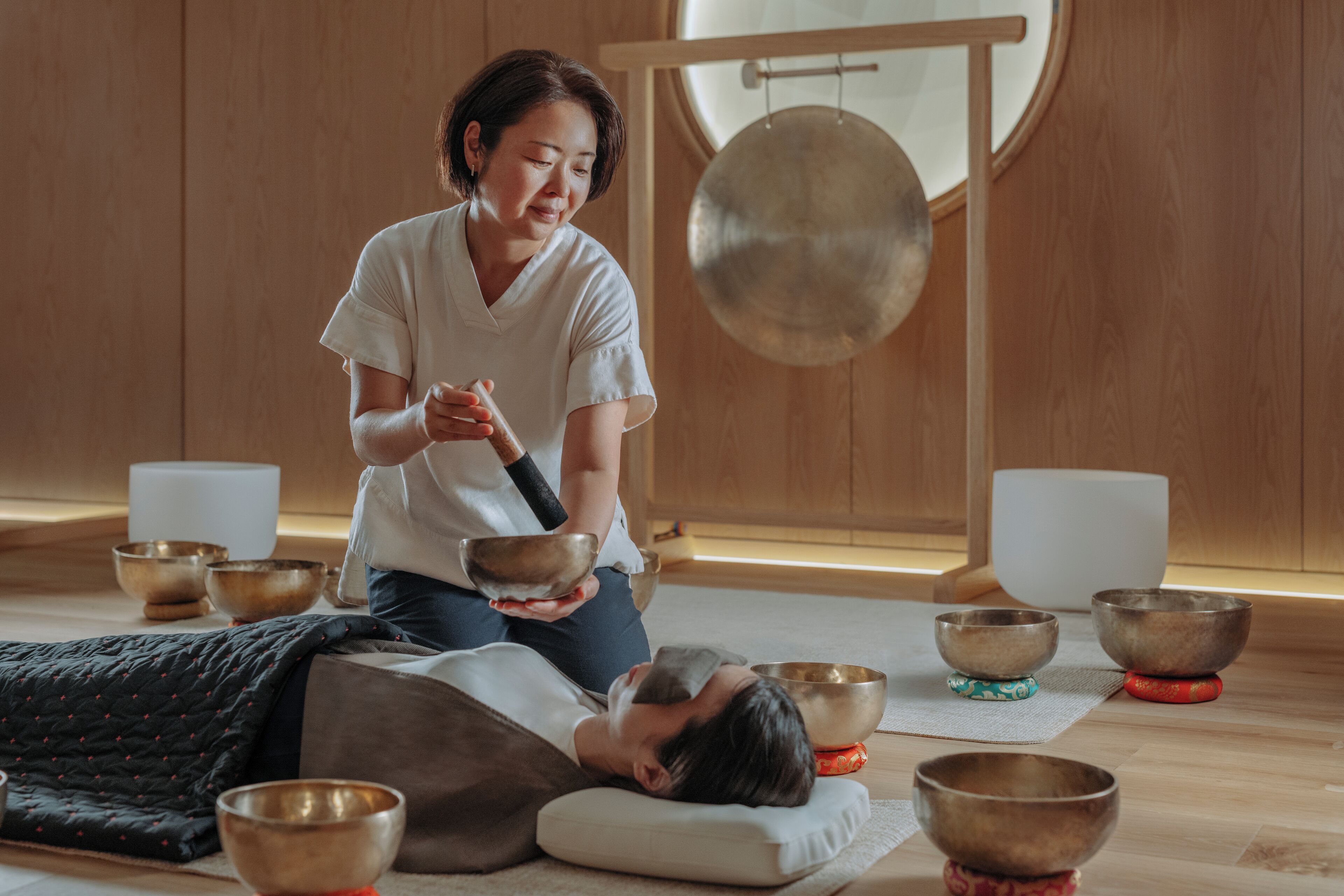 A room with a person lying on a mat, surrounded by several bowls and a gong, suggesting a meditation or wellness setting.