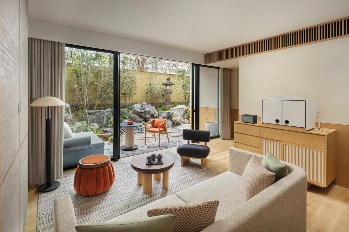 A modern living room with a sofa, coffee table, and chairs, a small table with a lamp, and a view of a garden with a stone lantern.
