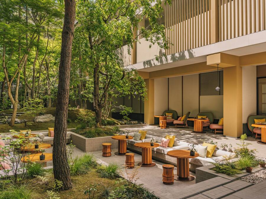 A modern outdoor seating area with wooden tables and chairs, surrounded by greenery and trees.