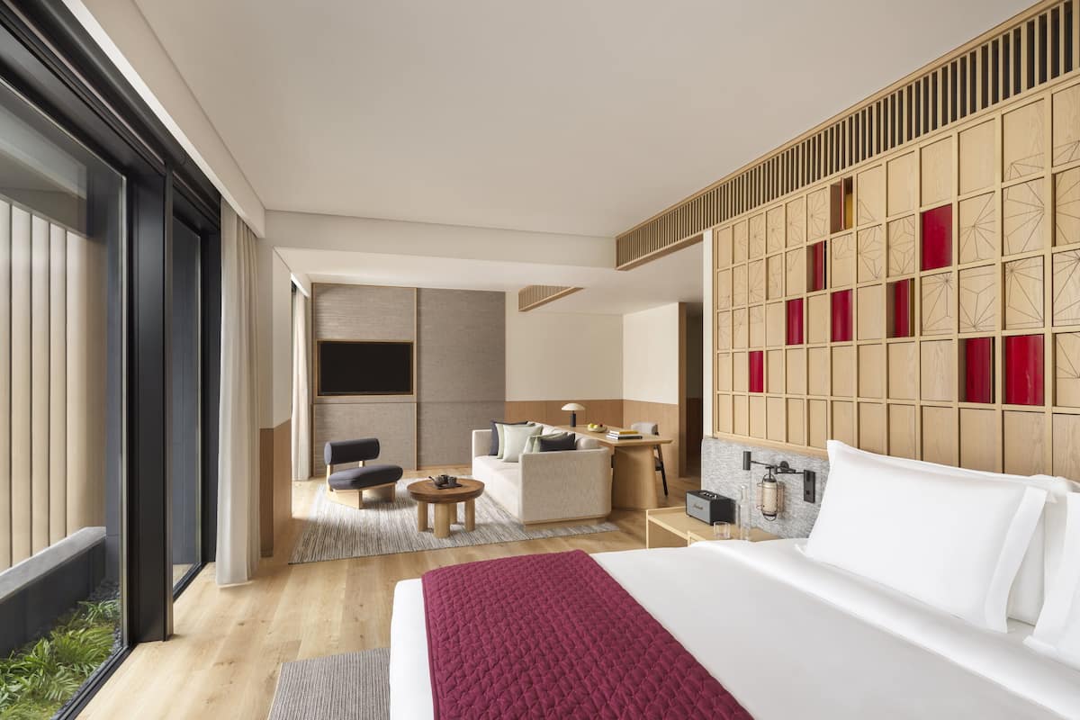 A modern hotel room with a large bed, a seating area with a sofa and chairs, a small table, and a view through a large window.