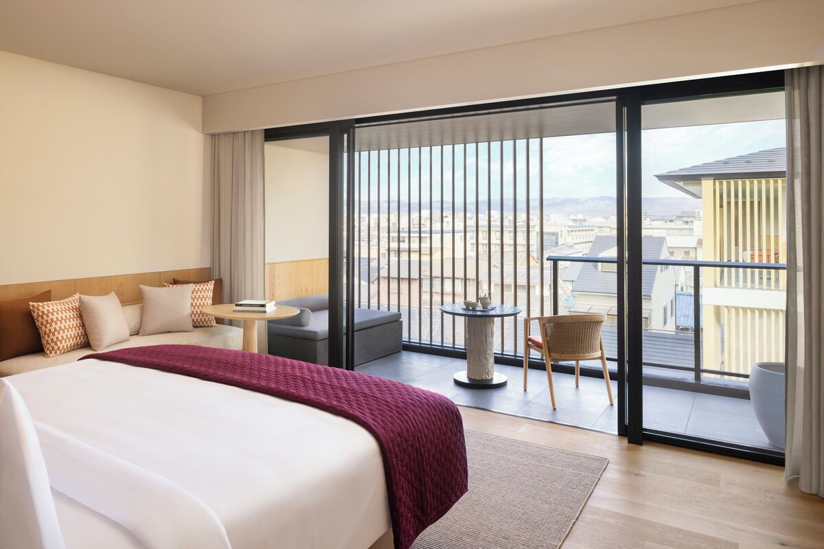 A hotel room with a large bed, a balcony with a view of the city, a small round table, and a sofa.