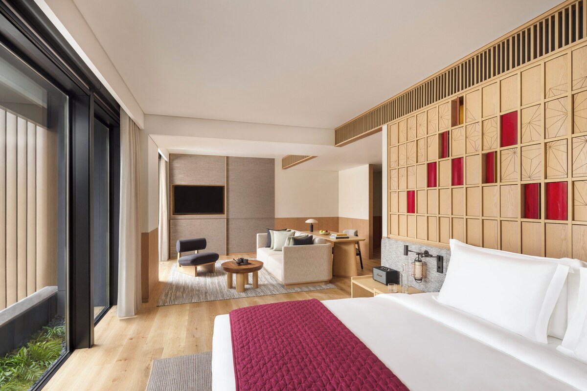 A modern hotel room with a large bed, a seating area with a sofa and chairs, a small table, and a view through a large window.