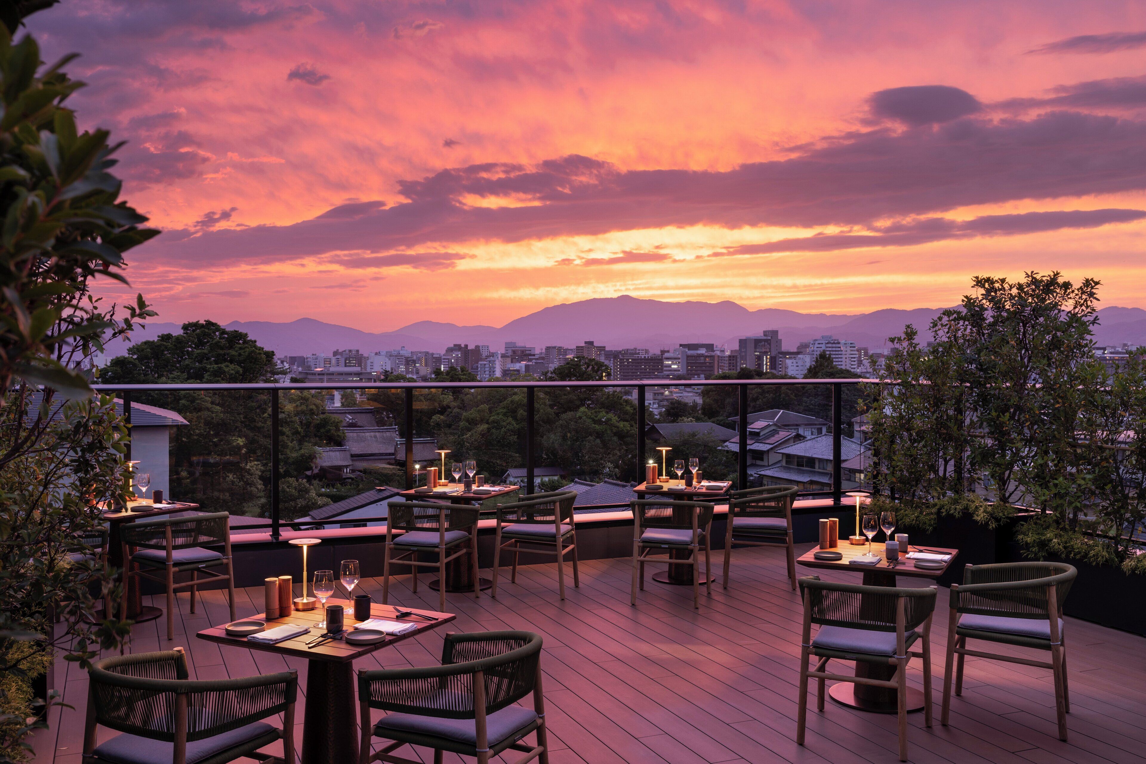 An outdoor dining area with tables and chairs, overlooking a cityscape and mountains during sunset.