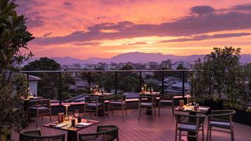 An outdoor dining area with tables and chairs, overlooking a cityscape and mountains during sunset.