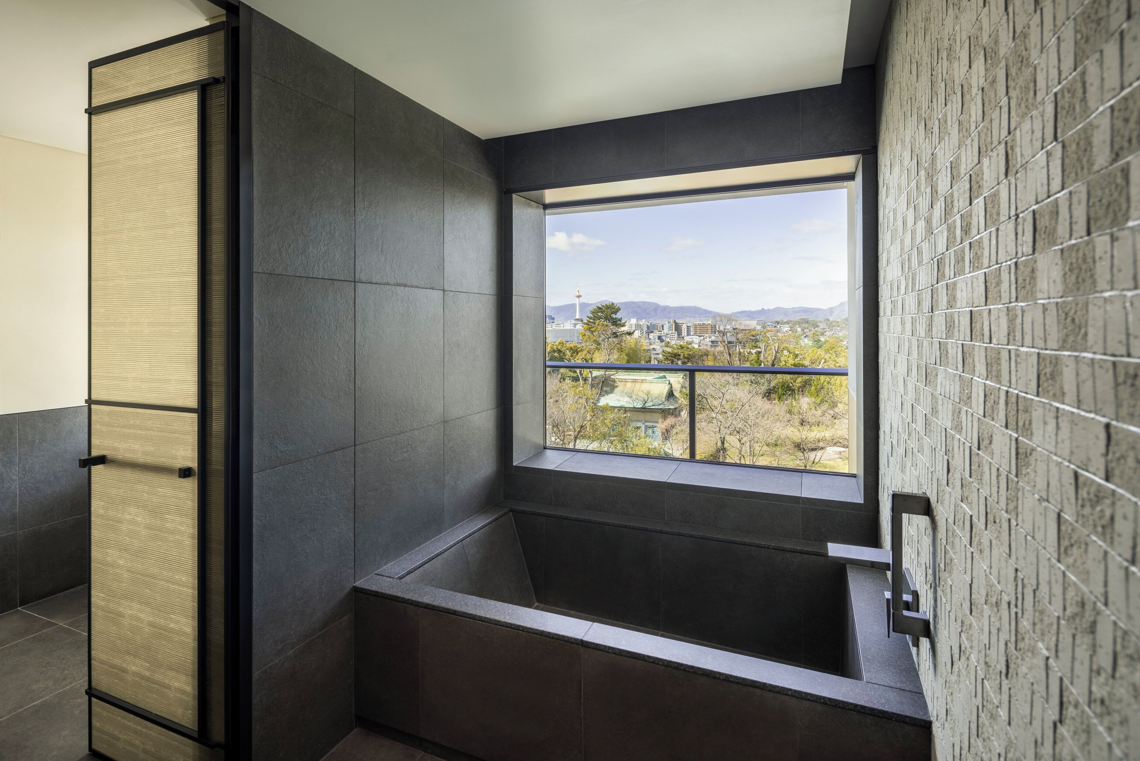 A modern bathroom with a large window, dark tiles, and a view of a cityscape.