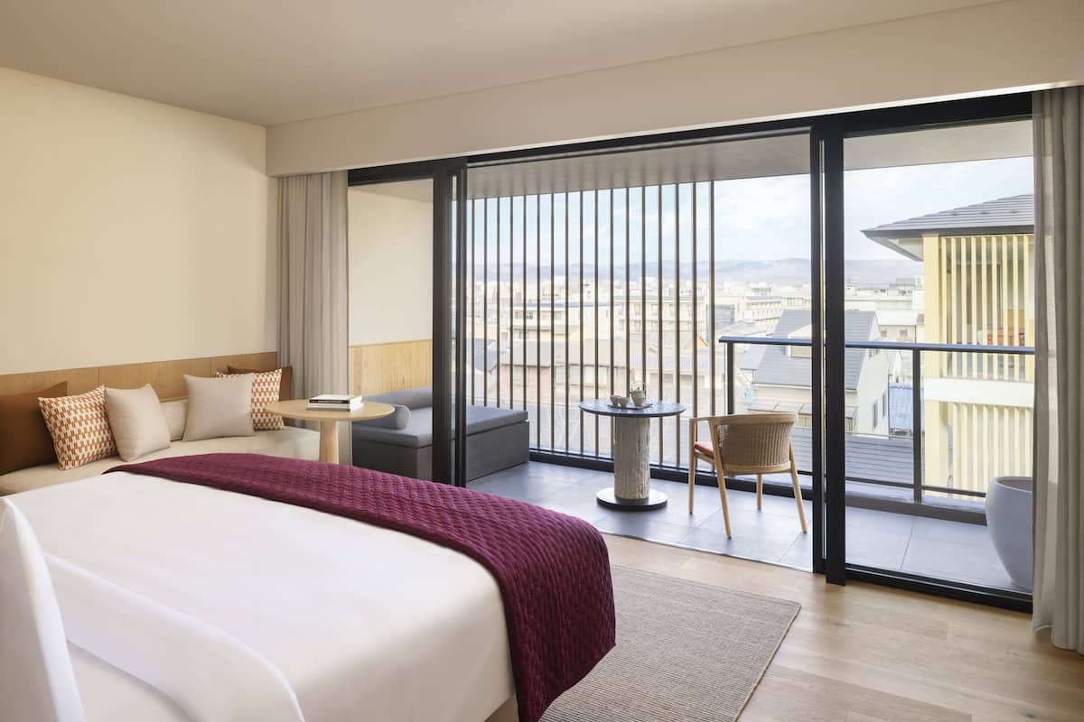 A hotel room with a large bed, a balcony with a view of the city, a small round table, and a sofa.