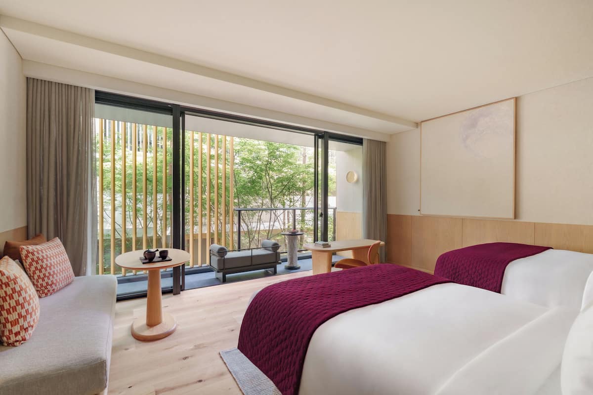 A hotel room with a large bed, a sofa, a small round table, and a balcony with a view of greenery.