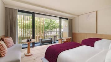 A hotel room with a large bed, a sofa, a small round table, and a balcony with a view of greenery.