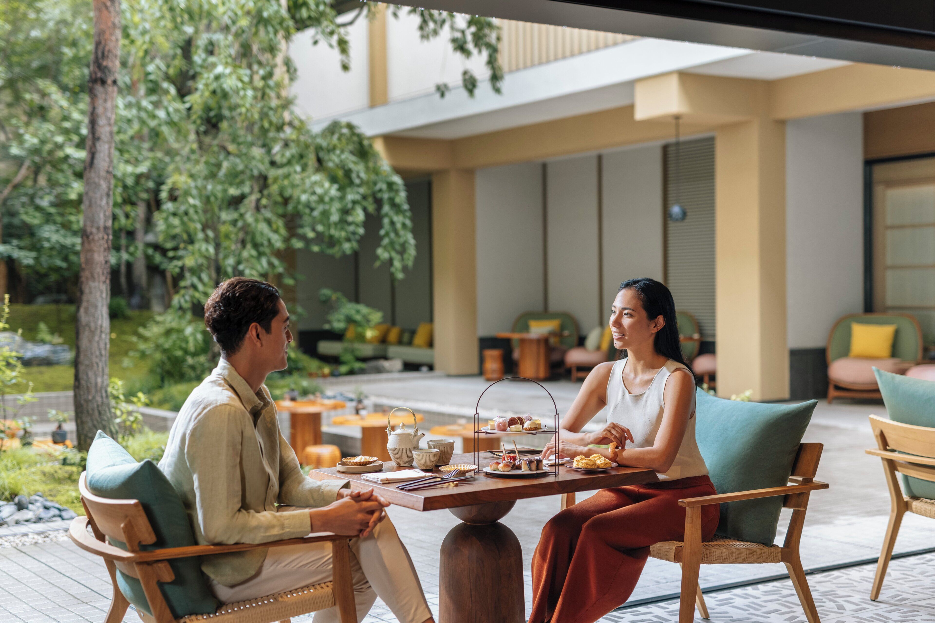 An outdoor seating area with a table set for two, surrounded by greenery and modern furniture.