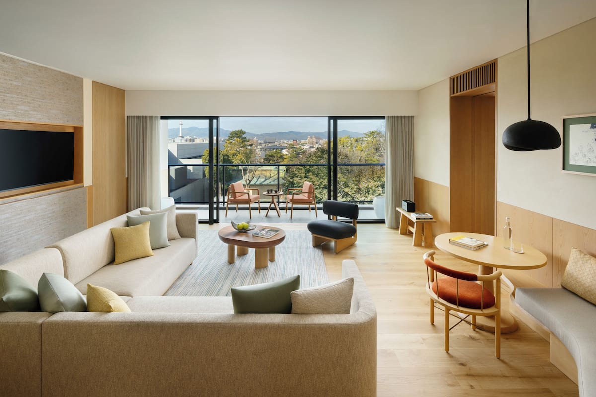 A modern living room with a sofa, chairs, a coffee table, and a dining table. There is a large window with a view of buildings and trees.