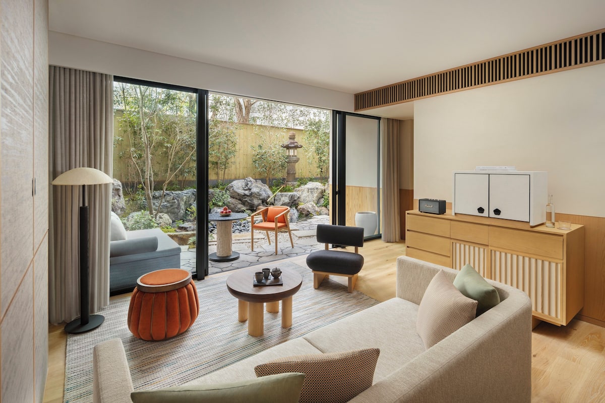 A modern living room with a sofa, coffee table, and chairs, a small table with a lamp, and a view of a garden with a stone lantern.
