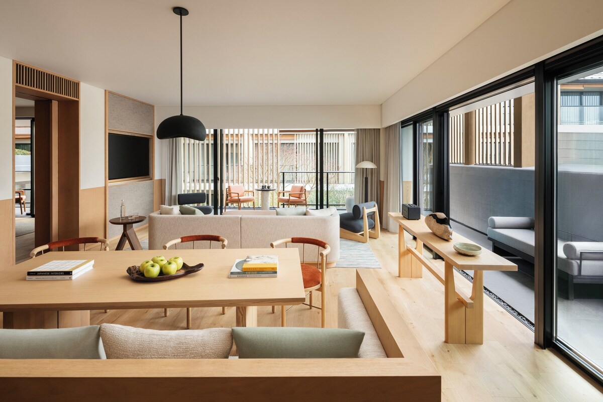 A modern living room with a sofa, dining table, and chairs. There is a large window with blinds, a hanging light fixture, and a view of the outside.
