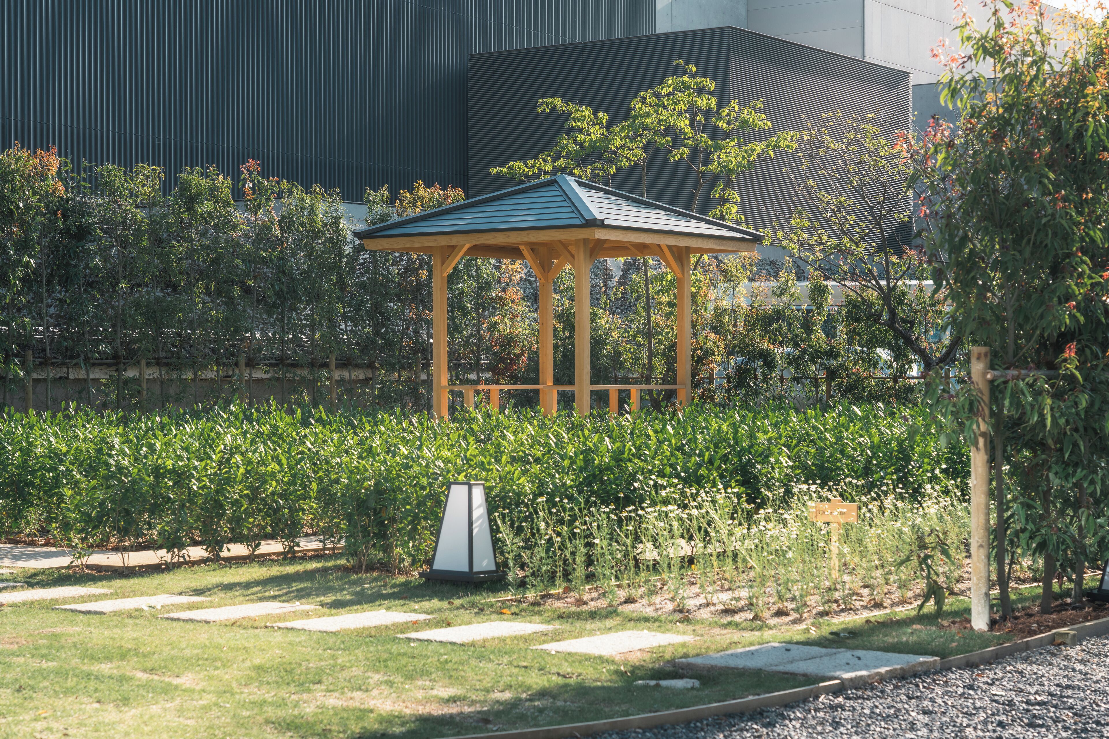 A gazebo with a green roof, surrounded by a garden with various plants and a gravel path.