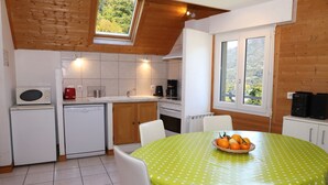 A compact kitchen with a dining area, featuring a green tablecloth, white chairs, and a fruit basket. - Appartement Les Pics (Luz-Saint-Sauveur)