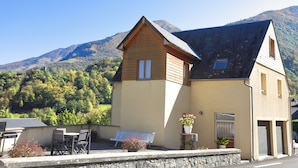 A two-story building with a sloped roof, a patio with a table and chairs, and a potted plant. - Appartement Les Pics (Luz-Saint-Sauveur)