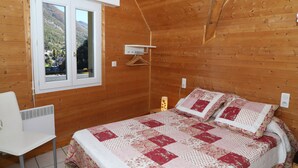 A wooden cabin bedroom with a bed, a chair, a window, and a hanging light fixture. - Appartement Les Pics (Luz-Saint-Sauveur)