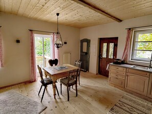 A cozy kitchen with wooden floors, a dining table, chairs, a hanging light fixture, and a window with curtains.