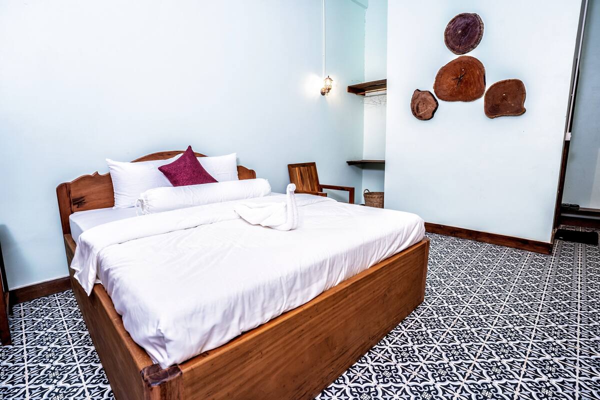 A neatly made bed with white linens and a wooden headboard, a wooden chair, and a wall with a light fixture and wooden wall art.