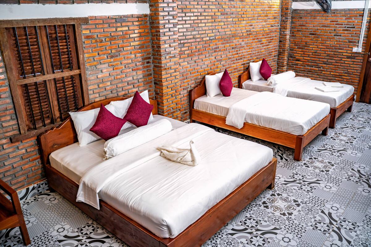 A hotel room with three wooden beds, each with white bedding and red and white pillows. The room features a brick wall and patterned floor tiles.