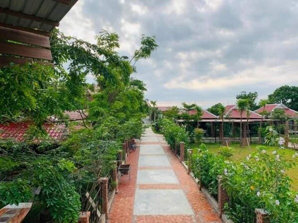 A pathway leading to a row of traditional-style buildings with red roofs, surrounded by lush greenery and a cloudy sky.