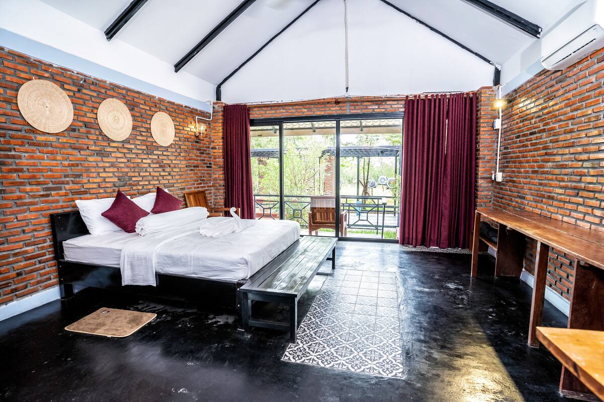 A room with a brick wall, a bed with white bedding, a bench, and a table.