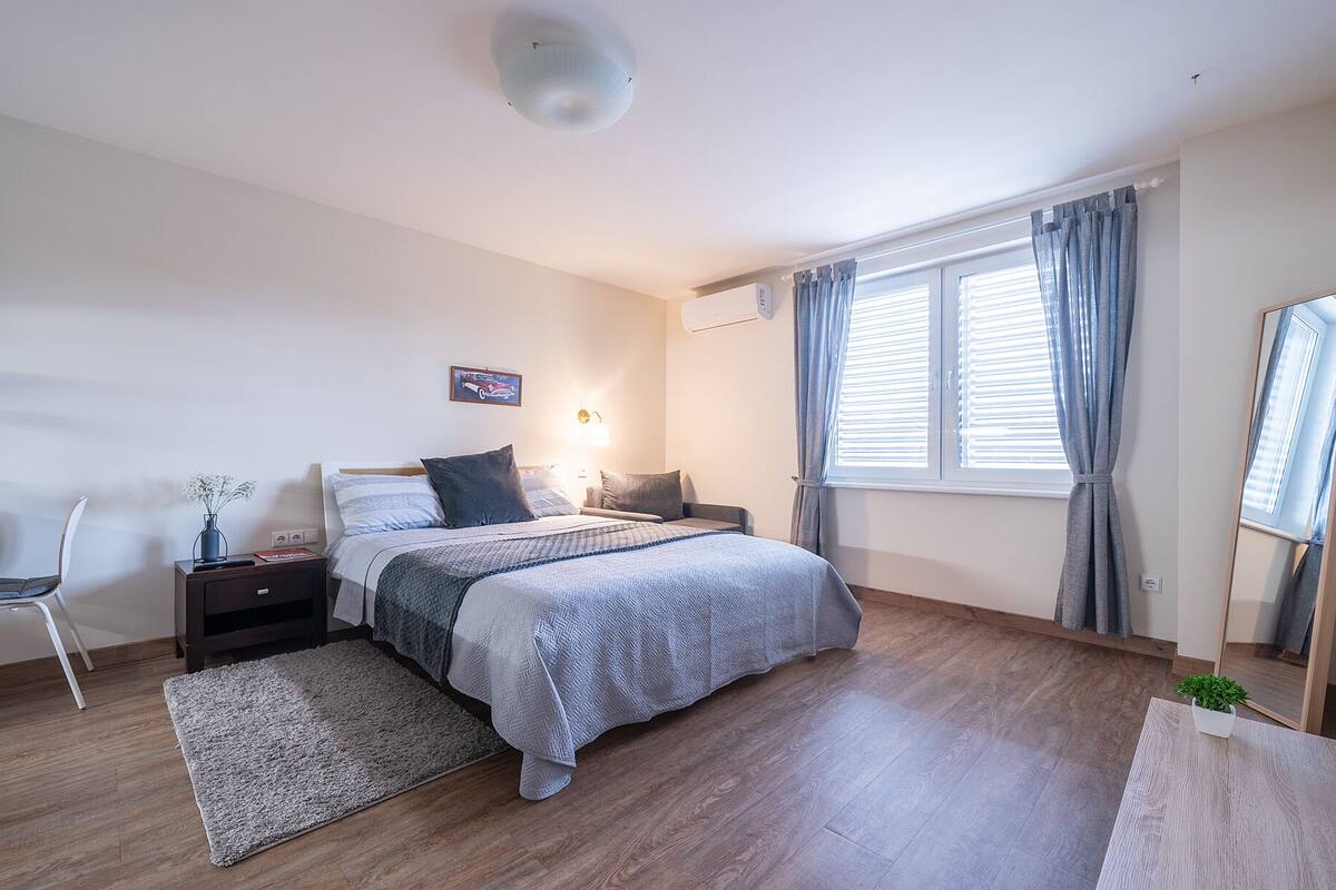 Ein Schlafzimmer mit Bett, Nachttischen, Schreibtisch, Stuhl, Teppich, einem Fenster mit Vorhängen und einer Deckenleuchte.