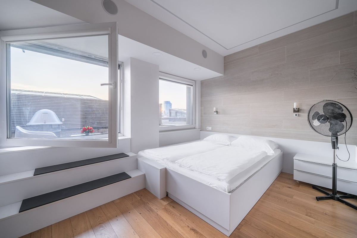 Ein modernes Schlafzimmer mit Bett, Standventilator und Fenster mit Blick auf die Stadt.