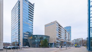 Una calle urbana moderna con edificios altos, incluyendo una estructura de vidrio curva y un edificio rectangular con una franja azul.