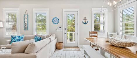 A modern living room with a beige sofa, a wooden dining table, and a bench. There are white walls, a door with a glass panel, and a view of greenery outside.
