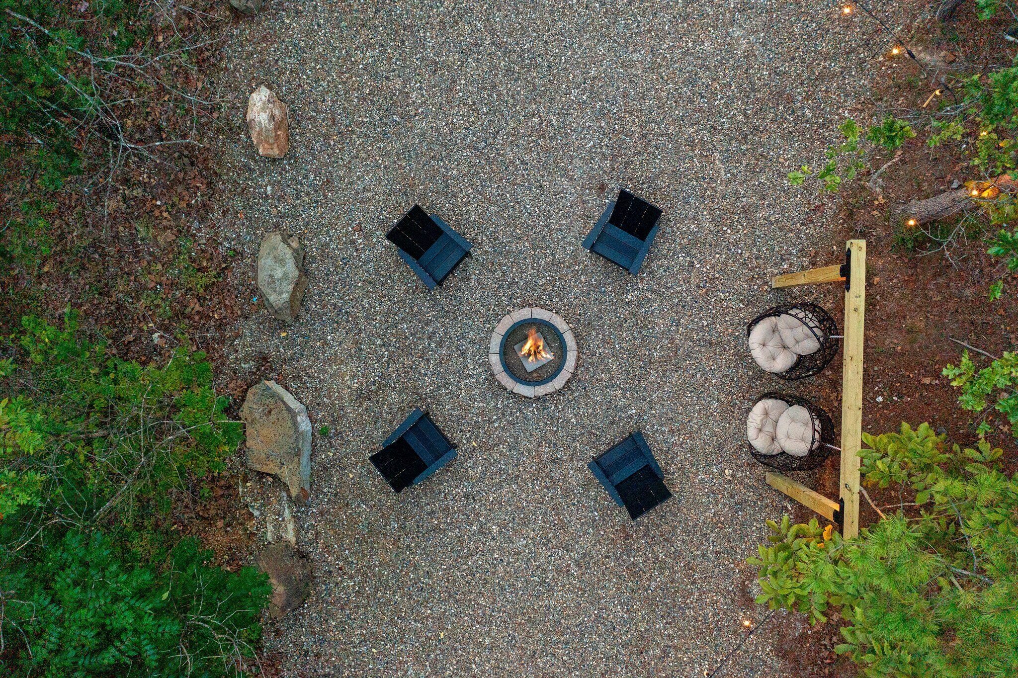 A circular outdoor seating area with a central fire pit, surrounded by six square seating cushions on a gravel surface.