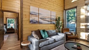 A cozy cabin interior with a wooden floor, a grey sofa with cushions, a small round table, and a painting on the wall.