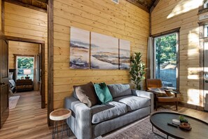 Un interior de cabaña acogedor, con piso de madera, un sofá gris con cojines, una mesita redonda y un cuadro en la pared.